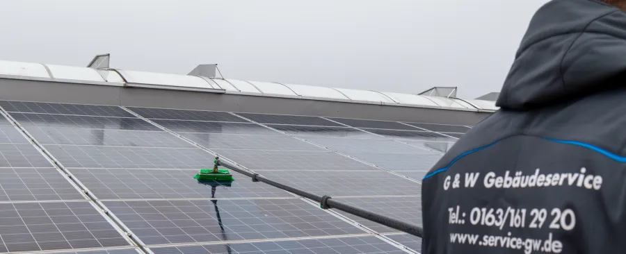 Ein G&W-Mitarbeiter in Firmenjacke reinigt mit der Teleskopbürste eine große Solaranlage auf einem Hallendach.