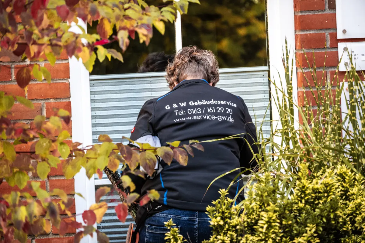 Eine Mitarbeiterin von G&W reinigt ein Fenster an einem Backsteinhaus, von hinten fotografiert, umgeben von Herbstlaub.